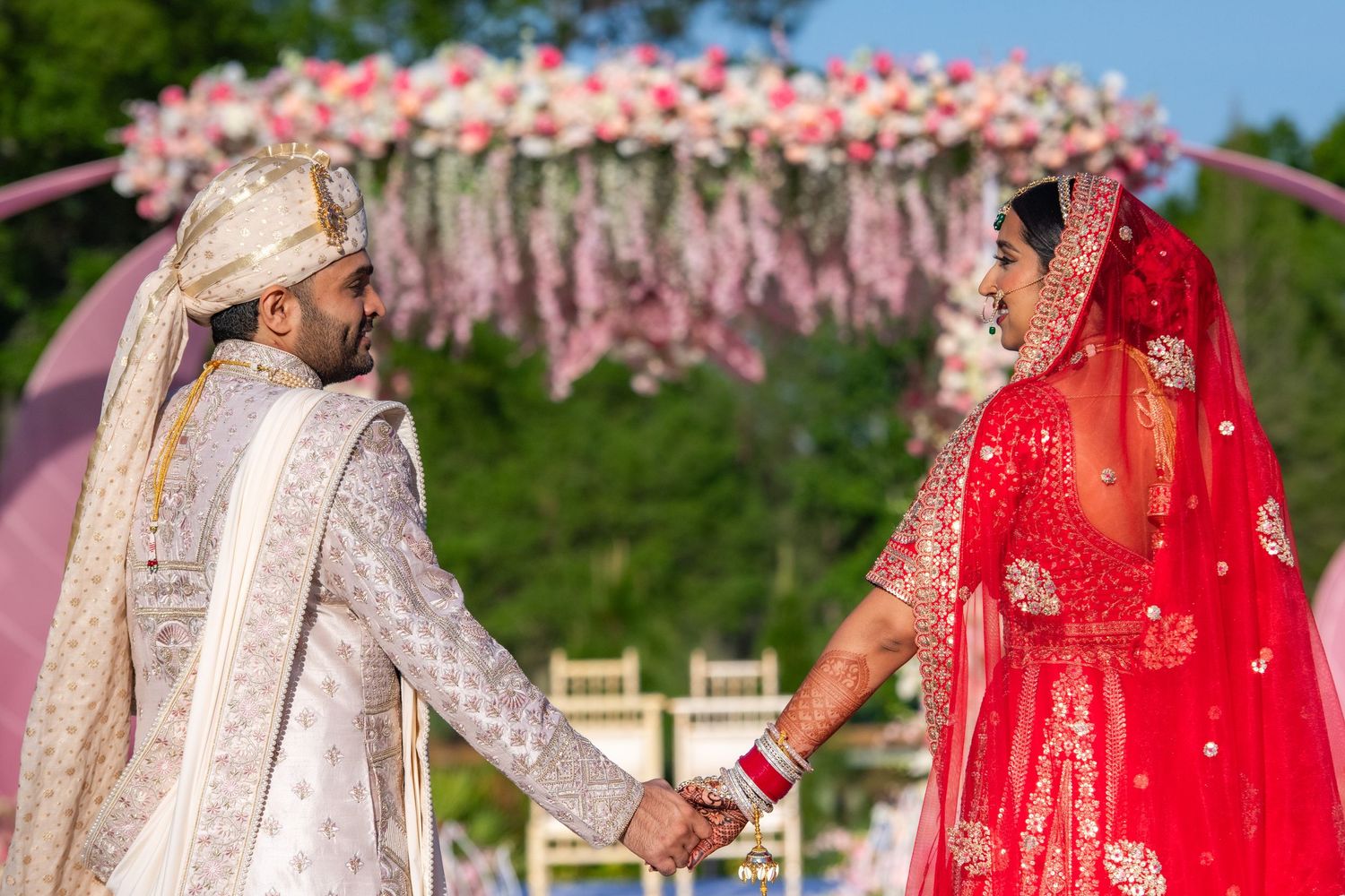 Couple in traditional Indian wedding attire holds hands under pink floral arch.