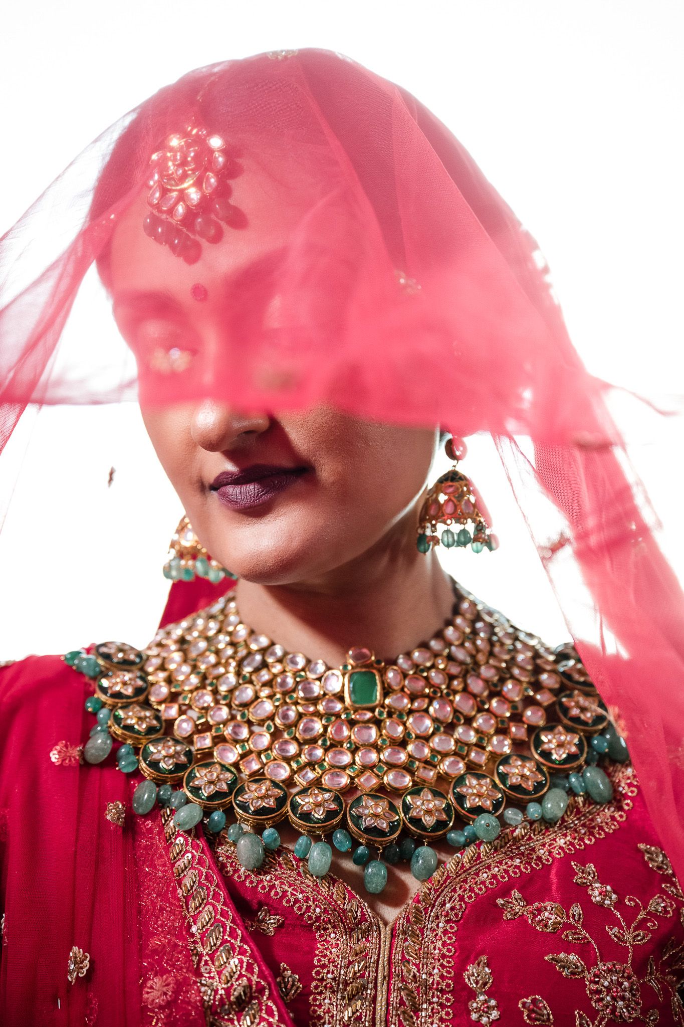 Traditional South Asian bridal jewelry featuring an ornate kundan necklace with emerald accents under a red veil.