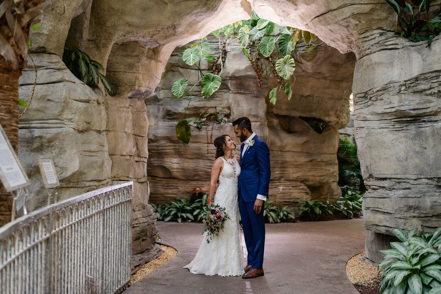 Wedding couple stands in natural stone grotto surrounded by ferns and romantic lighting.