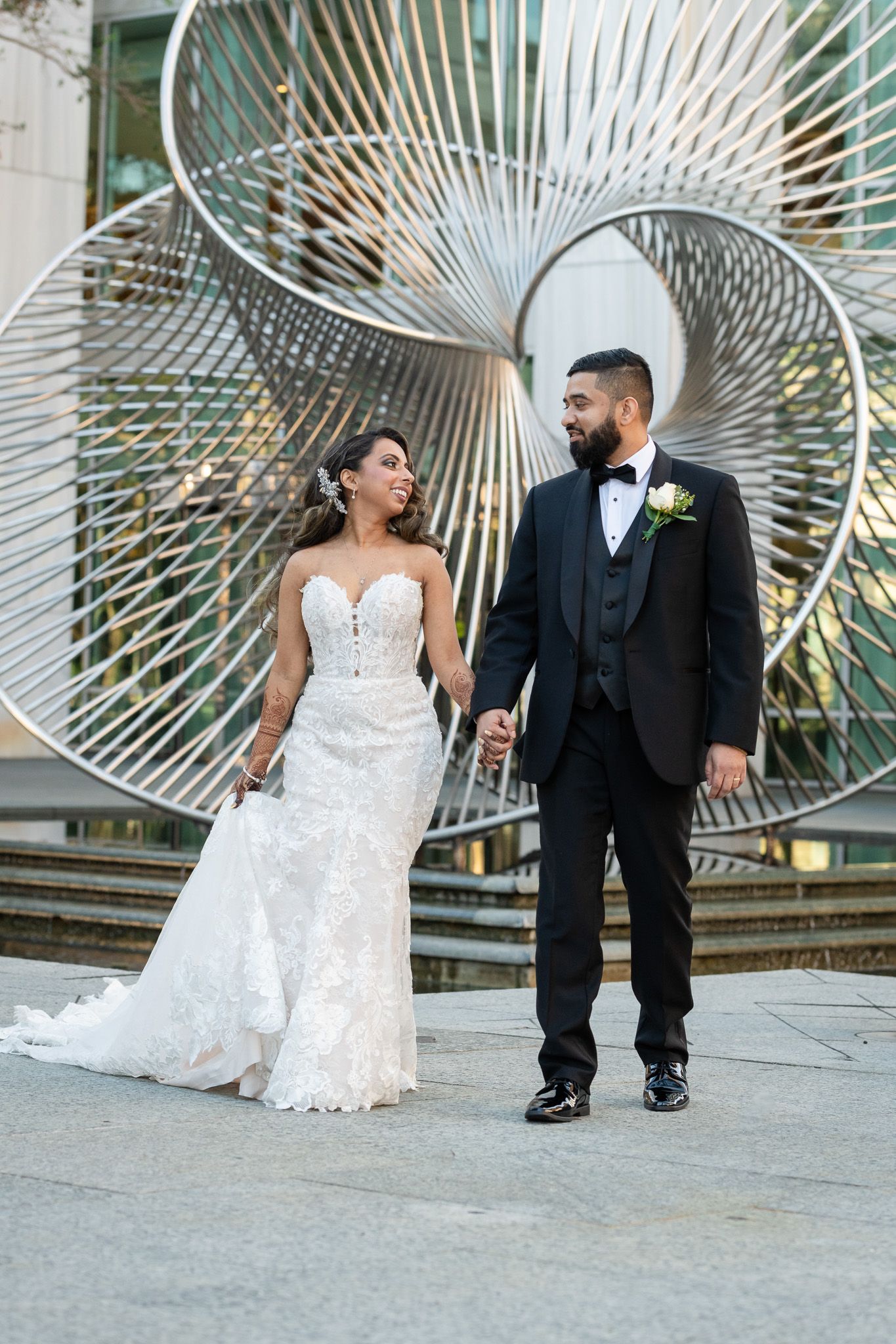 Couple poses in wedding attire in front of modern spiral metal art installation.