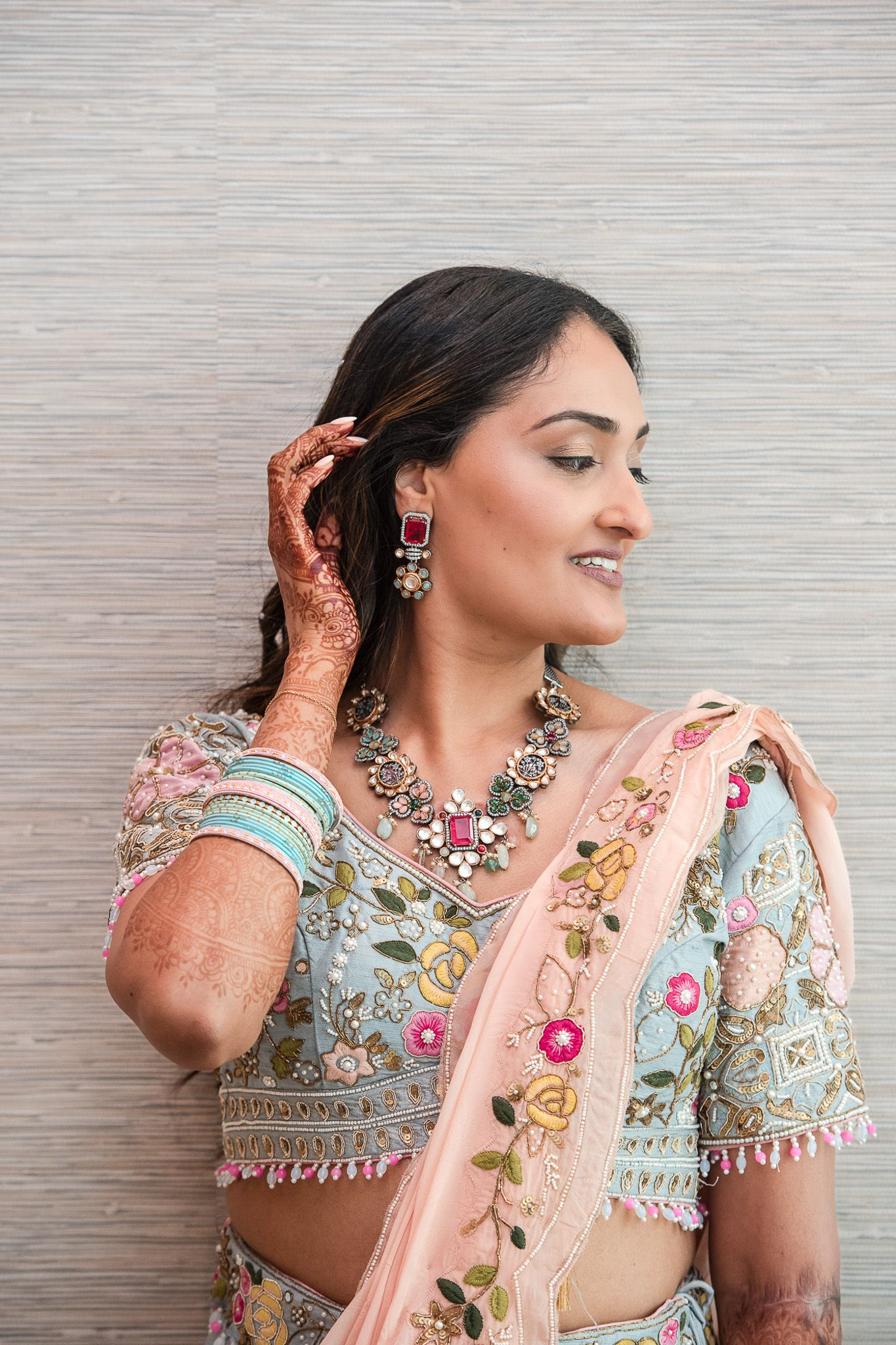 Bride in traditional South Asian attire with intricate mehndi designs and elegant jewelry poses against textured wall.