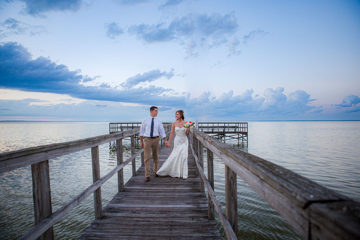 Sunset wedding portrait of couple standing on wooden pier over calm waters.