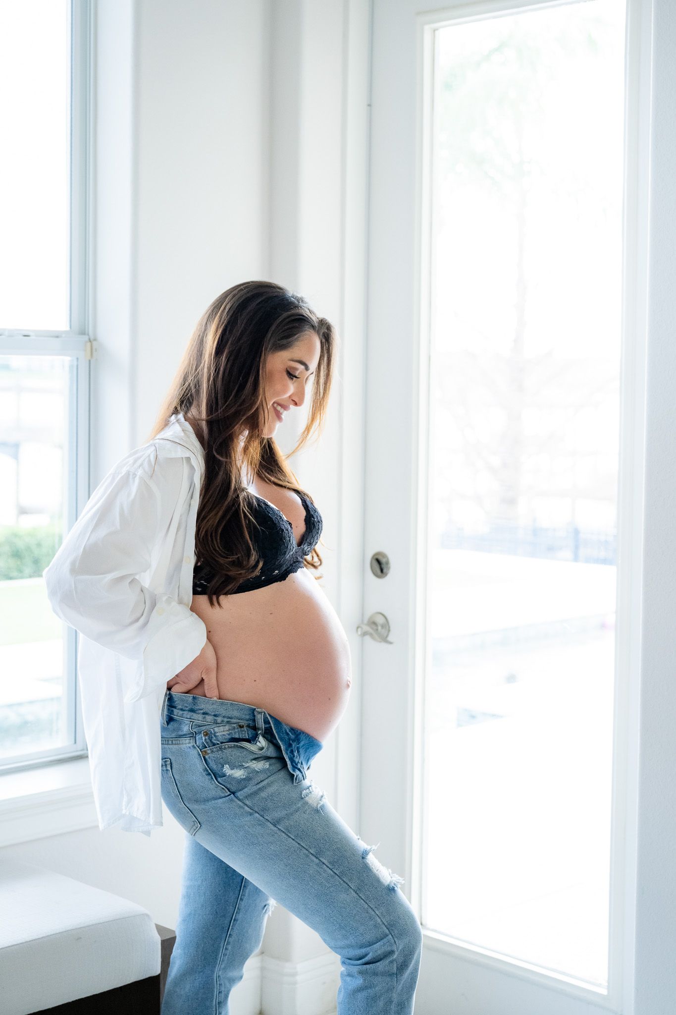 Expectant mother in white shirt and jeans poses by window in casual maternity photo.