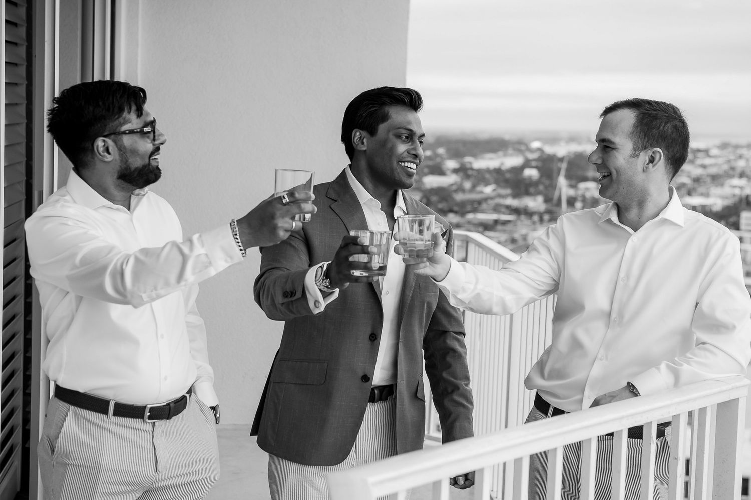Three people enjoying drinks and conversation on a balcony overlooking a coastal view.