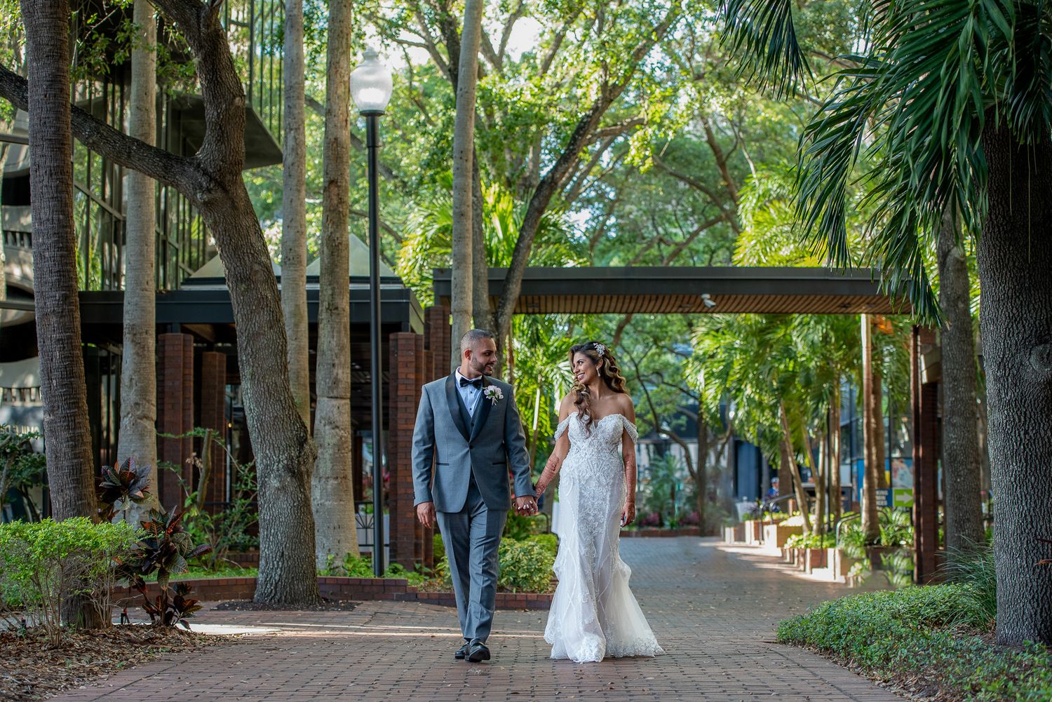 Newlyweds walking together along tree-lined path with tropical foliage.