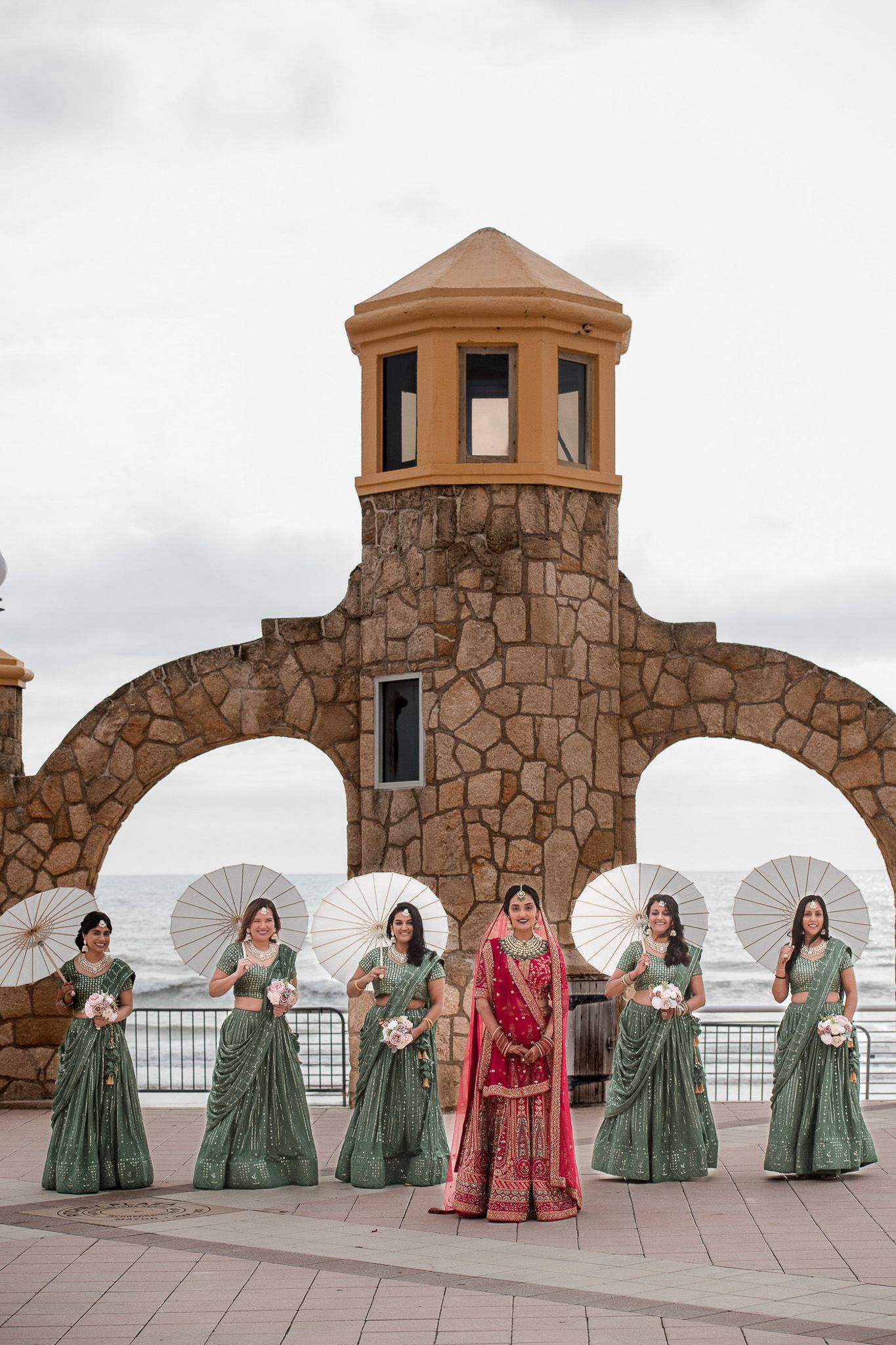 Bride in red lehenga surrounded by bridesmaids in green holding white parasols under stone archway.