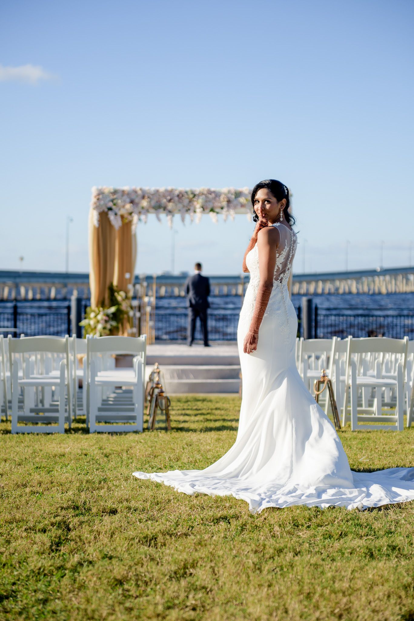 A bride in a white form-fitting gown poses on grass near a waterfront ceremony setup with floral arch.