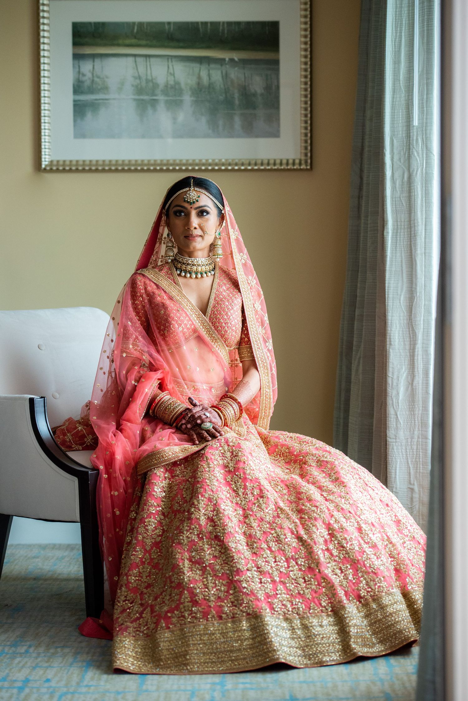 Traditional Indian bride in coral lehenga with gold embroidery poses elegantly by window.