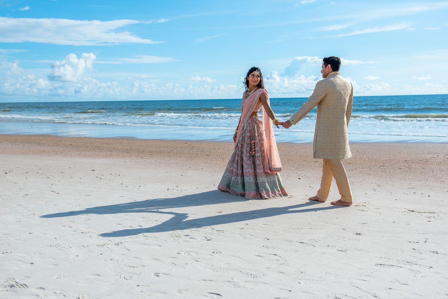 Couple in traditional Indian wedding attire holding hands on a scenic beach.