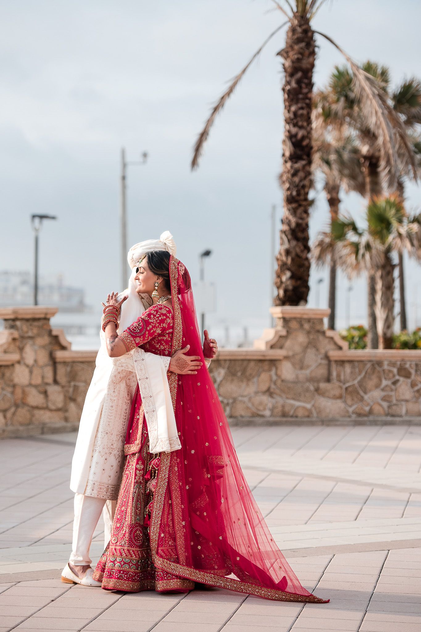 Couple in traditional South Asian wedding attire embracing on coastal pathway with palm trees.
