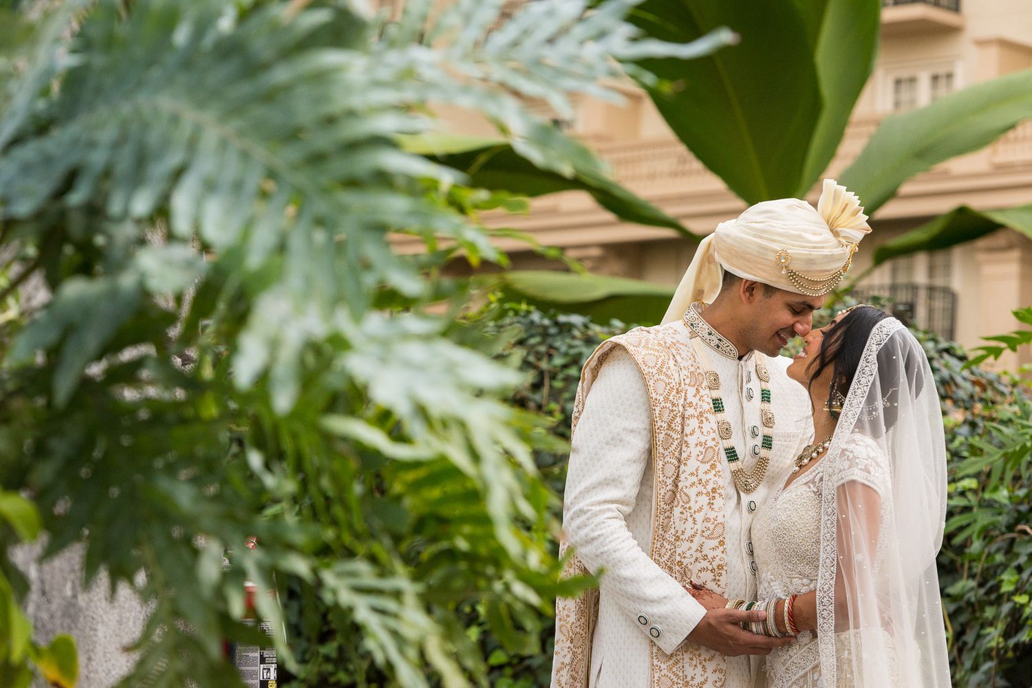 Newlyweds in traditional South Asian wedding attire embrace among lush greenery in a garden setting.