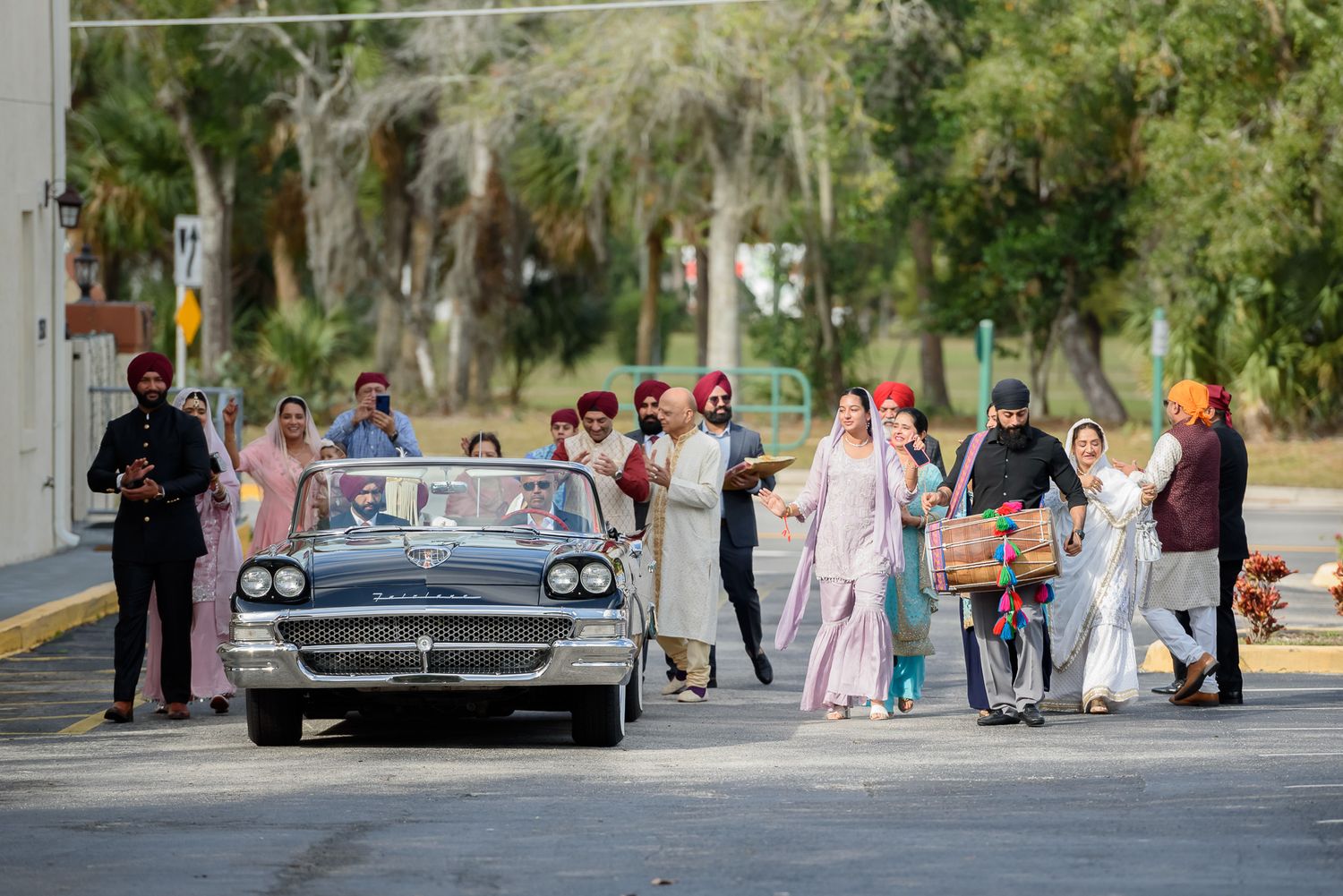 Vintage car procession with group in traditional attire on urban street.