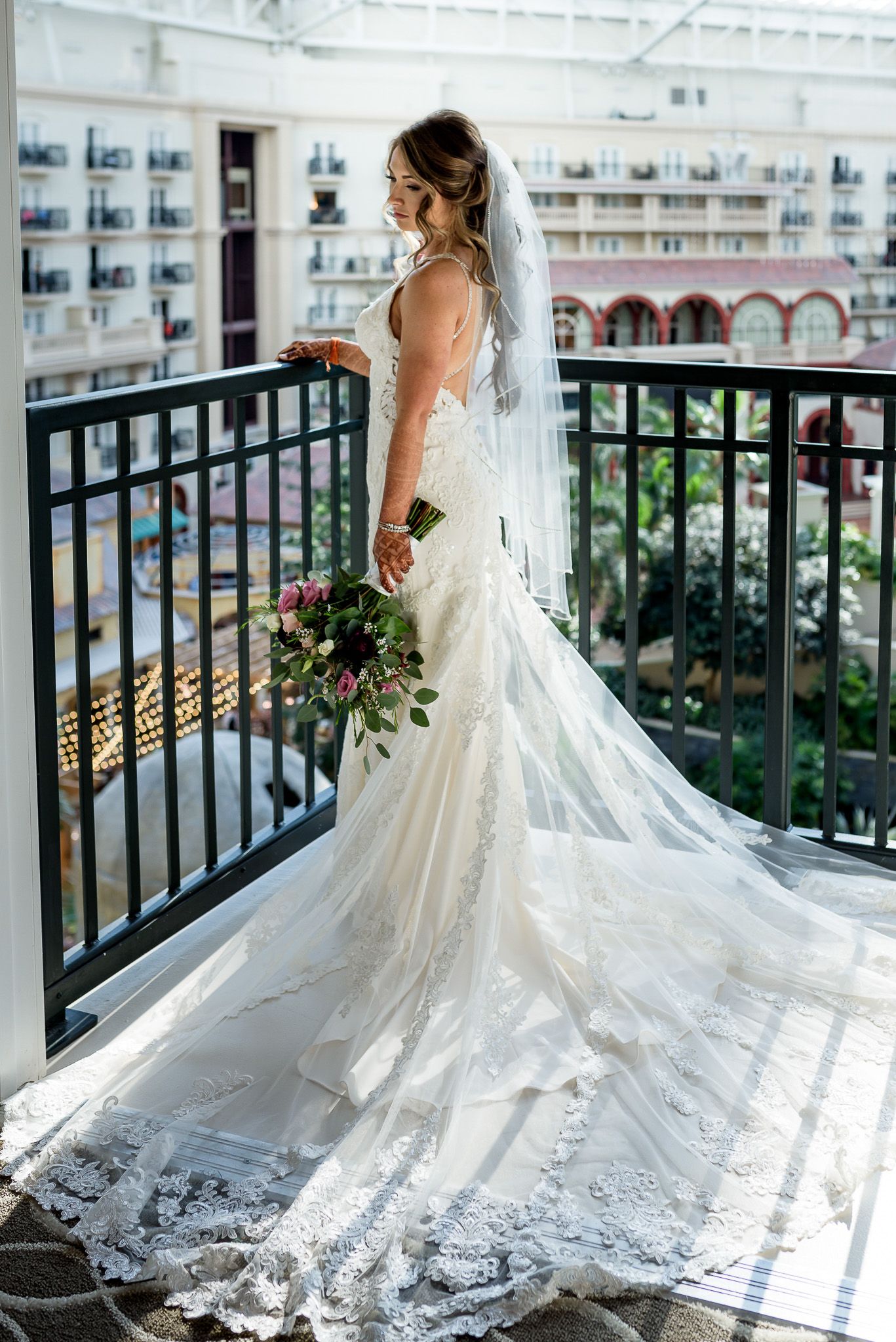 Bride in lace gown with flowing veil poses on balcony overlooking courtyard.