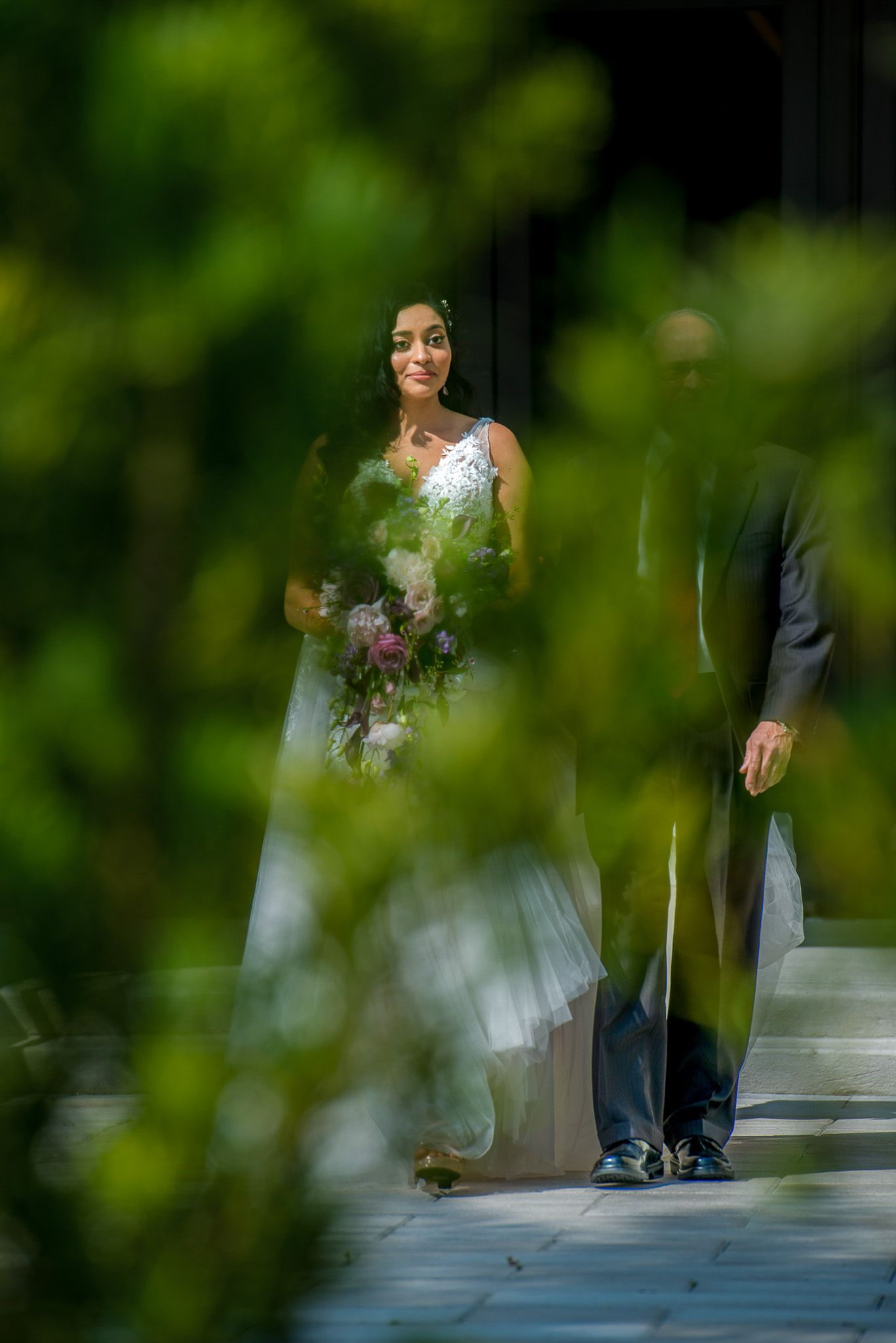 Creative wedding portrait through foliage showing bride in white dress on walkway.