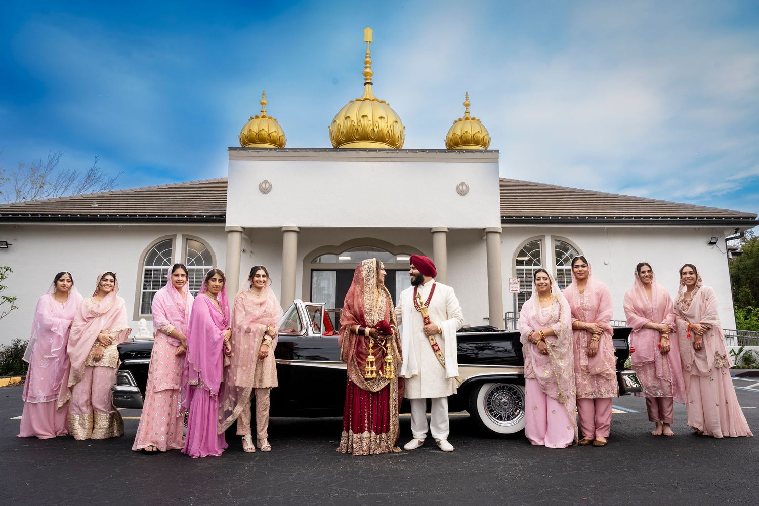 Wedding party gathered outside a Sikh temple with golden domes against blue sky.