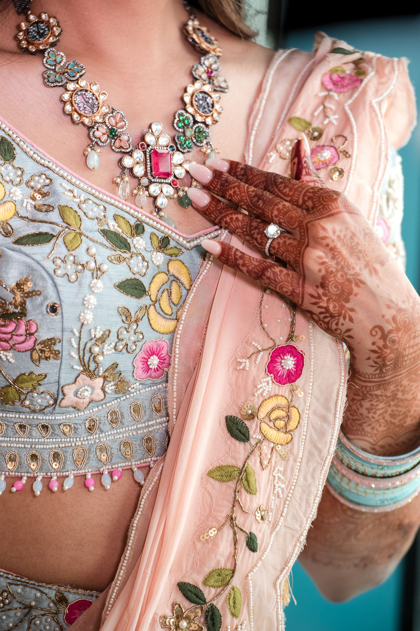 Detailed shot of ornate silver necklace and floral embroidered fabric with mehndi-adorned hands.