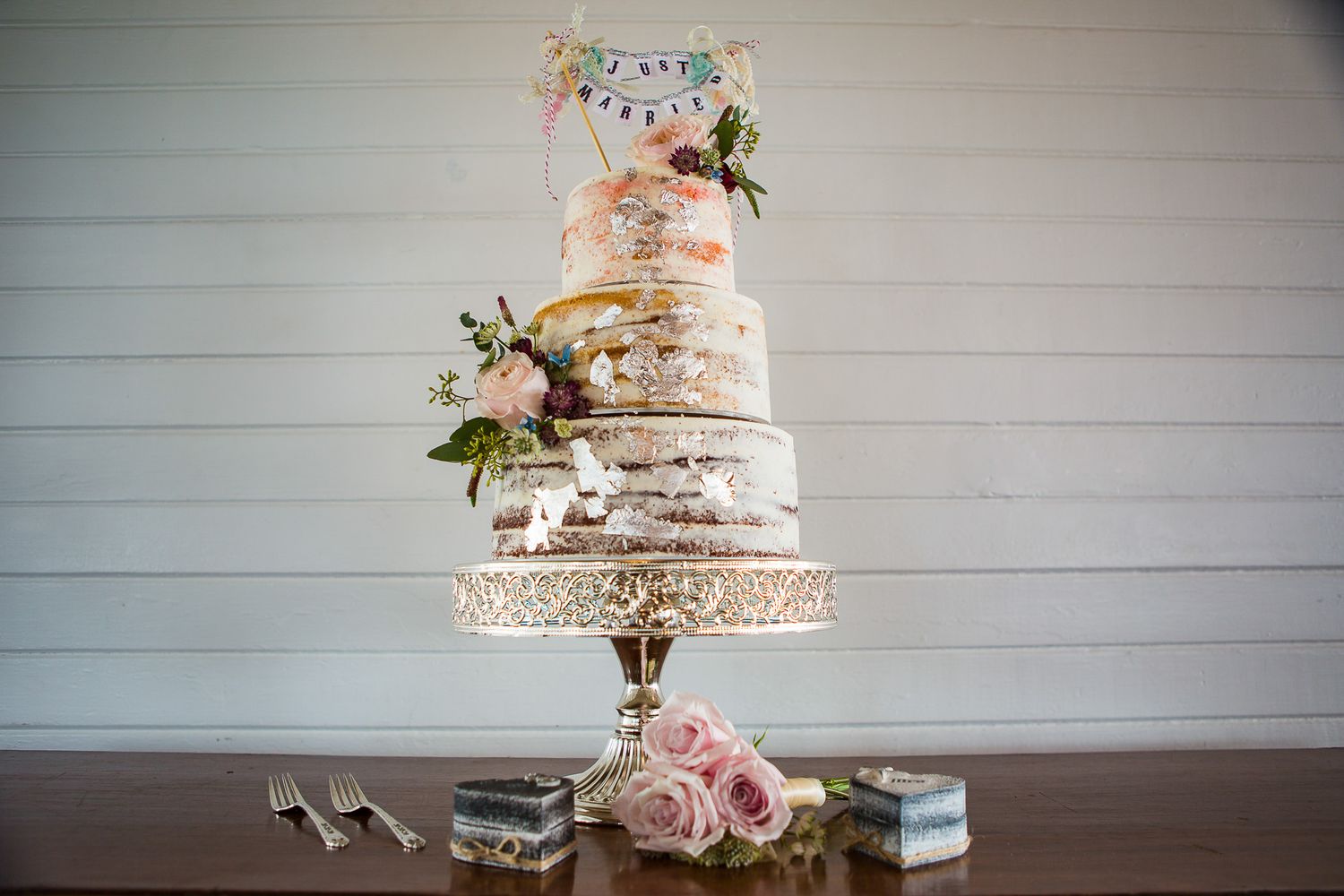 Elegant three-tier semi-naked wedding cake decorated with fresh pink roses and greenery on a wooden table.
