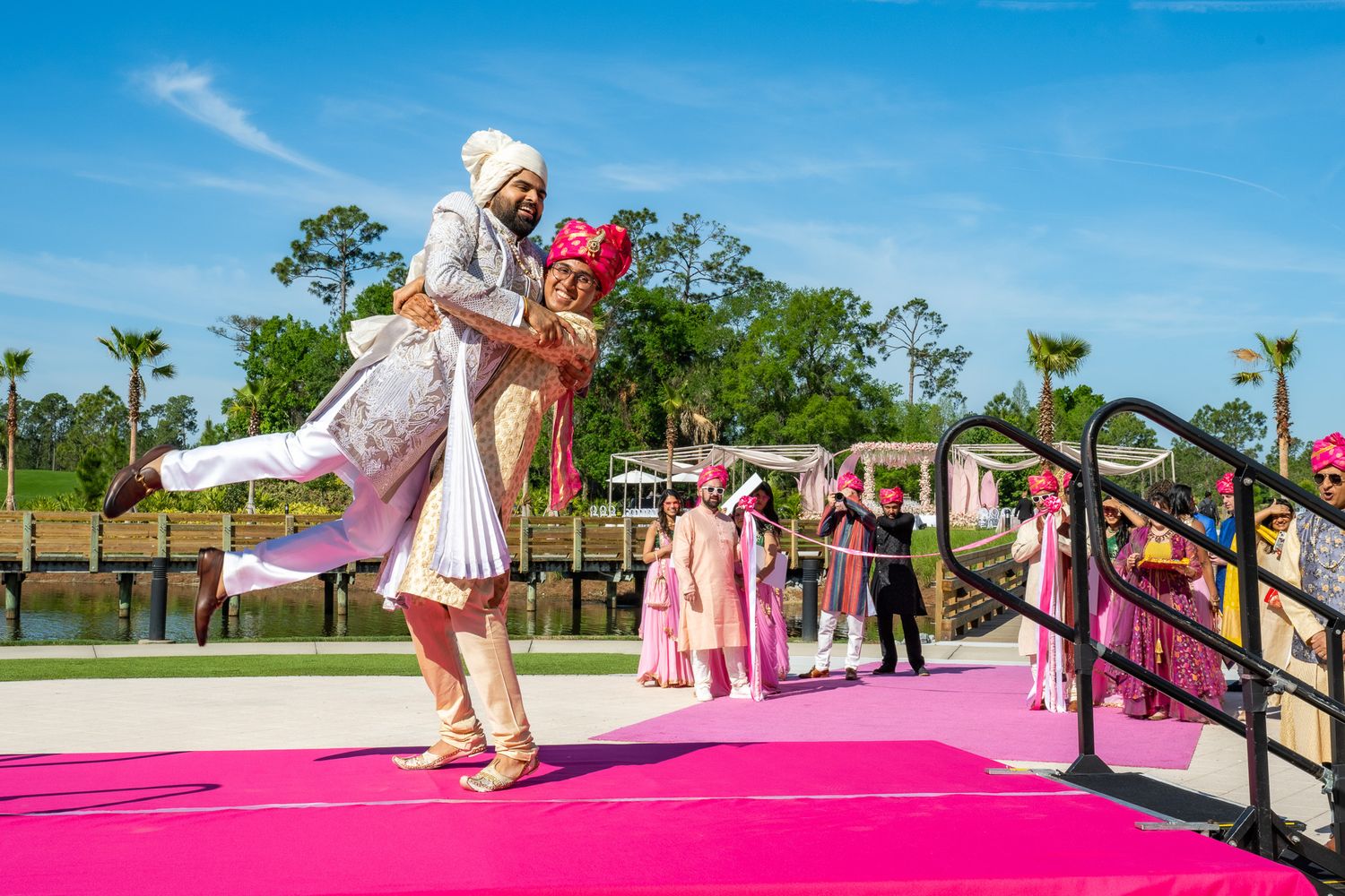 Groom in traditional attire performs dance moves on pink walkway during baraat ceremony.