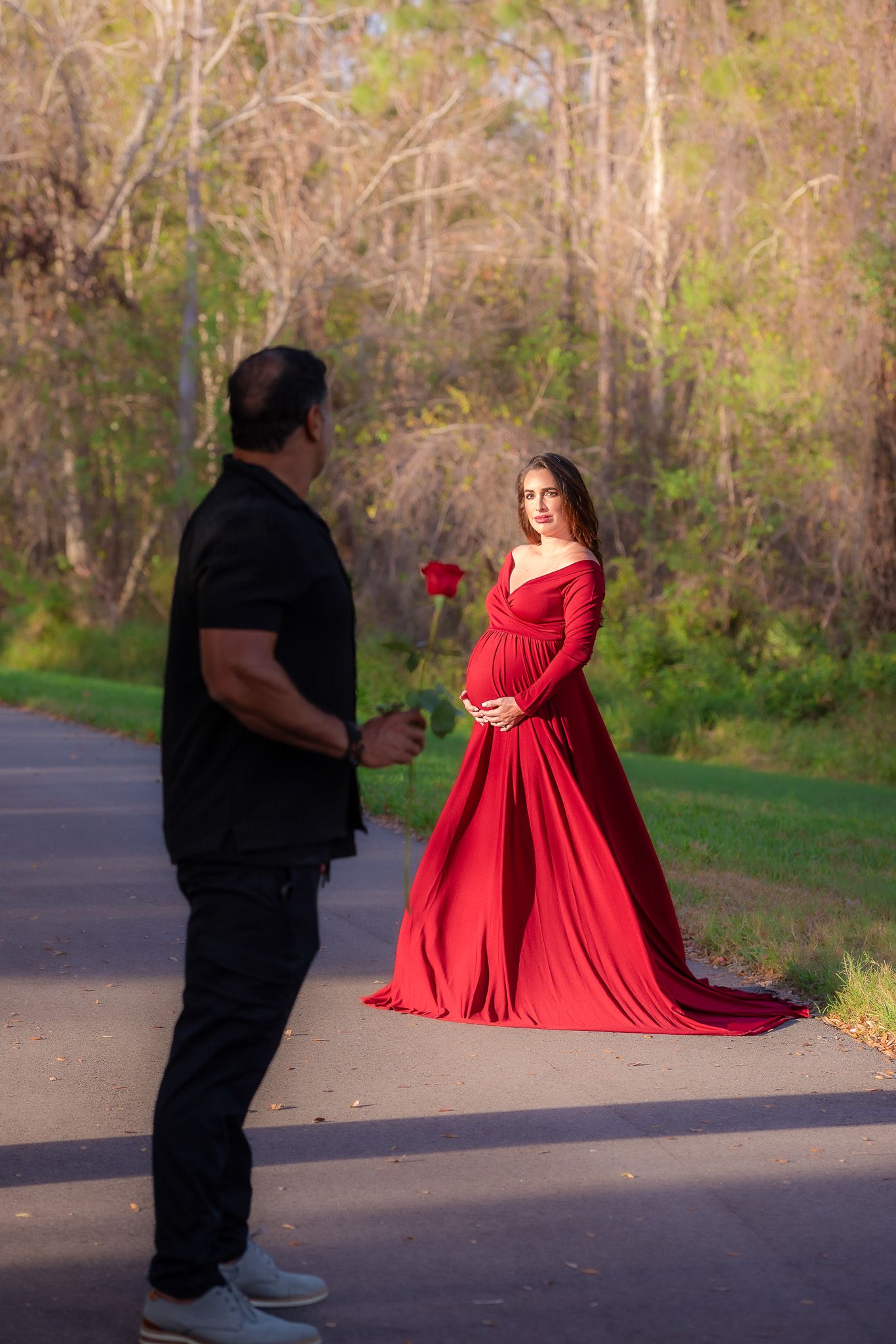 Maternity photo of woman in red gown while partner holds rose on wooded path.