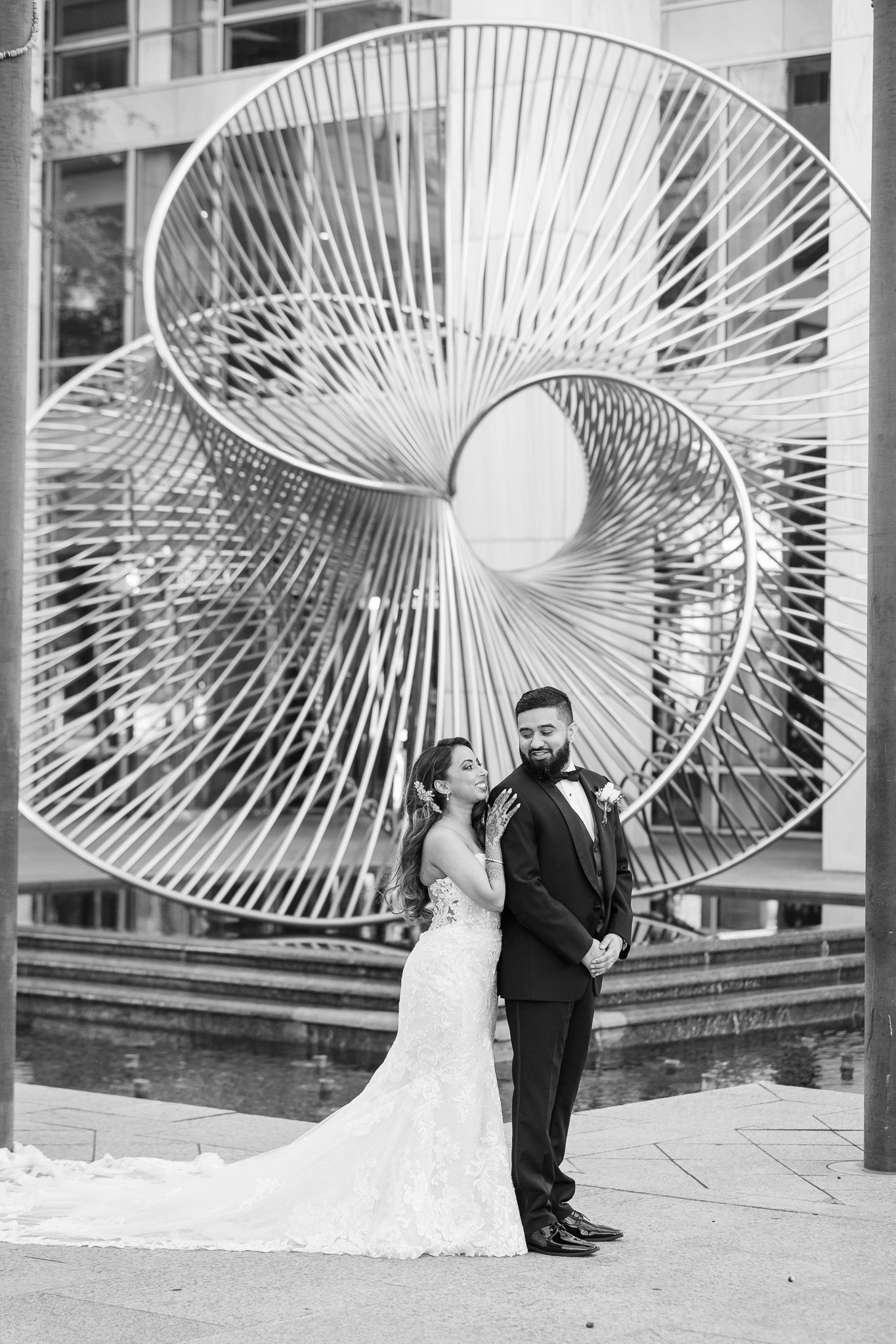A wedding couple poses in front of a large modern art sculpture with radiating metal spokes.