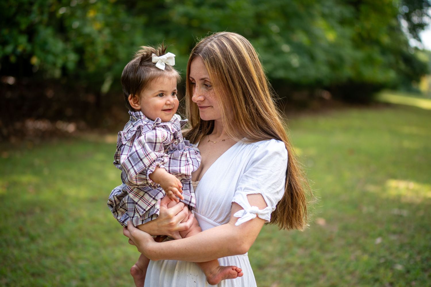 A mother in a white dress holds her young daughter in a floral outfit outdoors on a sunny day.