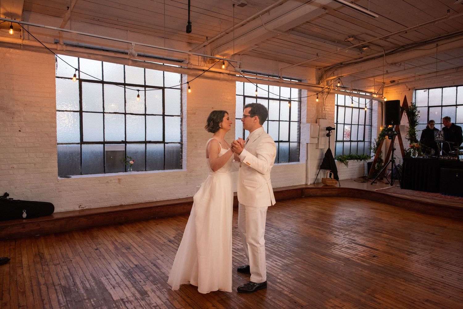 A newlywed couple dances together in a sunlit industrial venue with wooden floors.