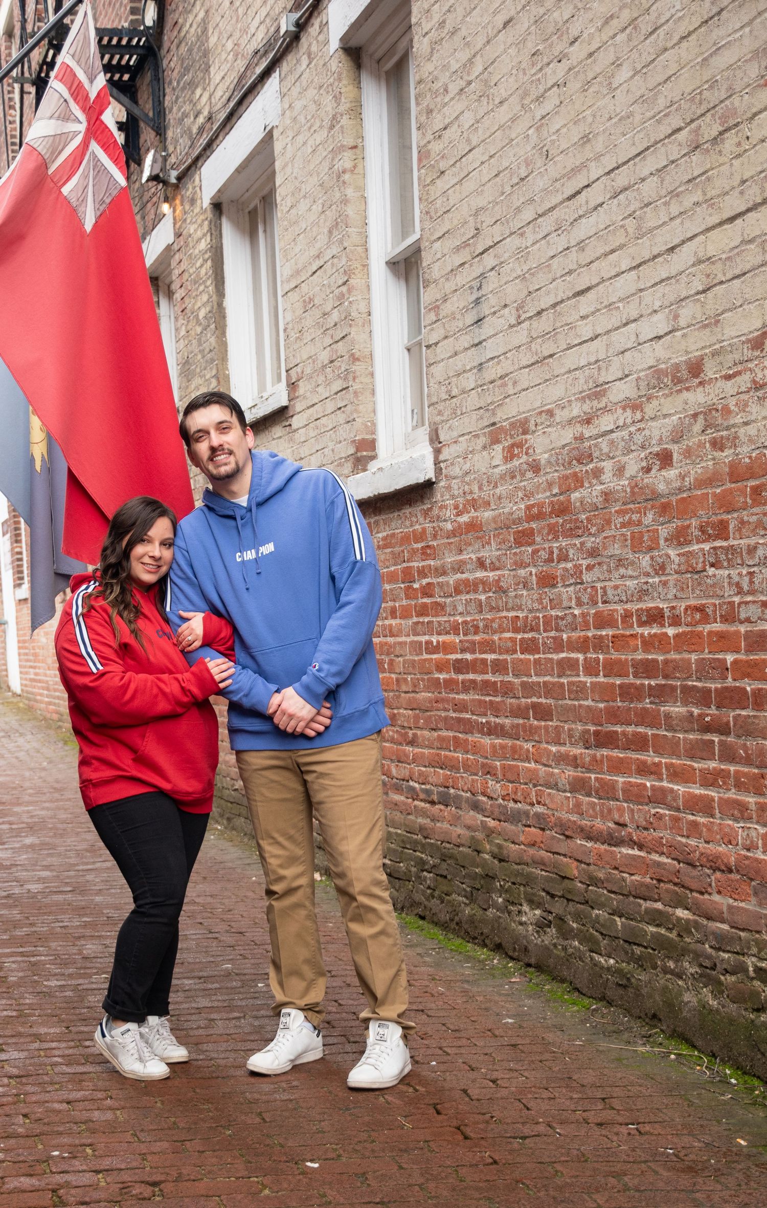 Couple in casual wear poses playfully against brick wall with flags.