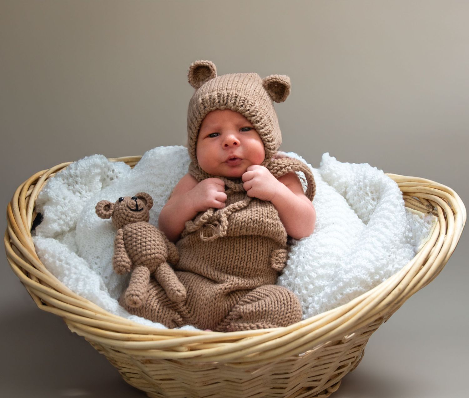 Baby wearing knit hat and sweater rests peacefully in woven basket with white blanket.