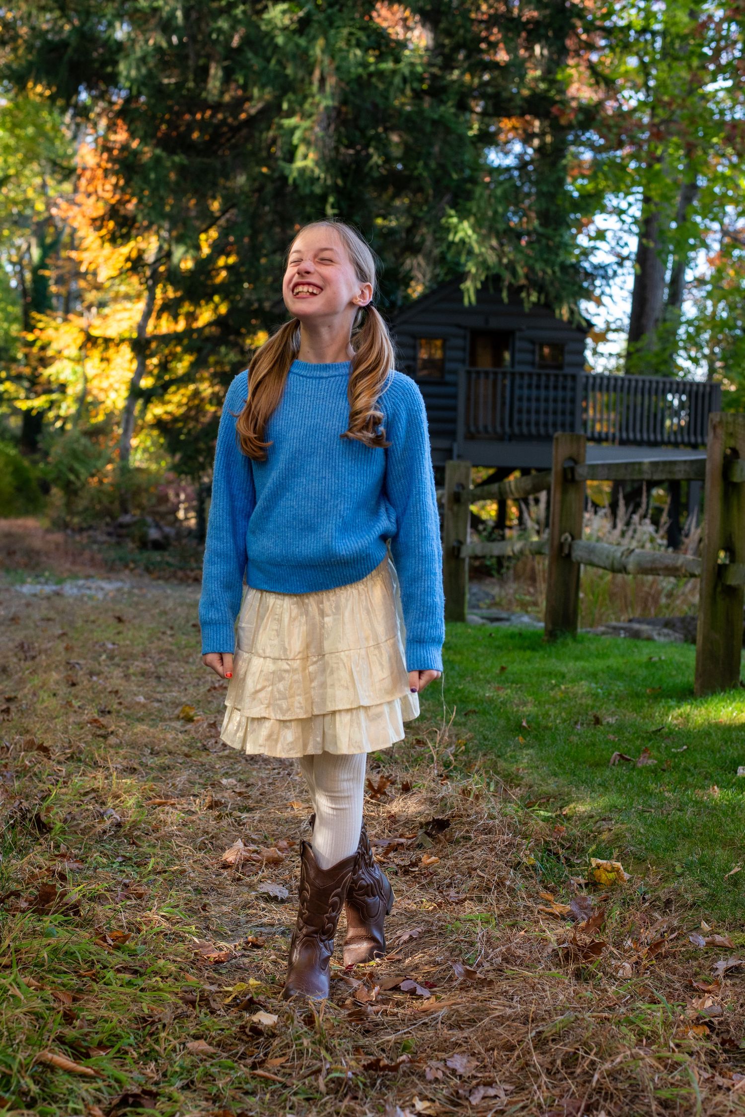 Girl in blue sweater and layered skirt smiling outdoors with treehouse in background.