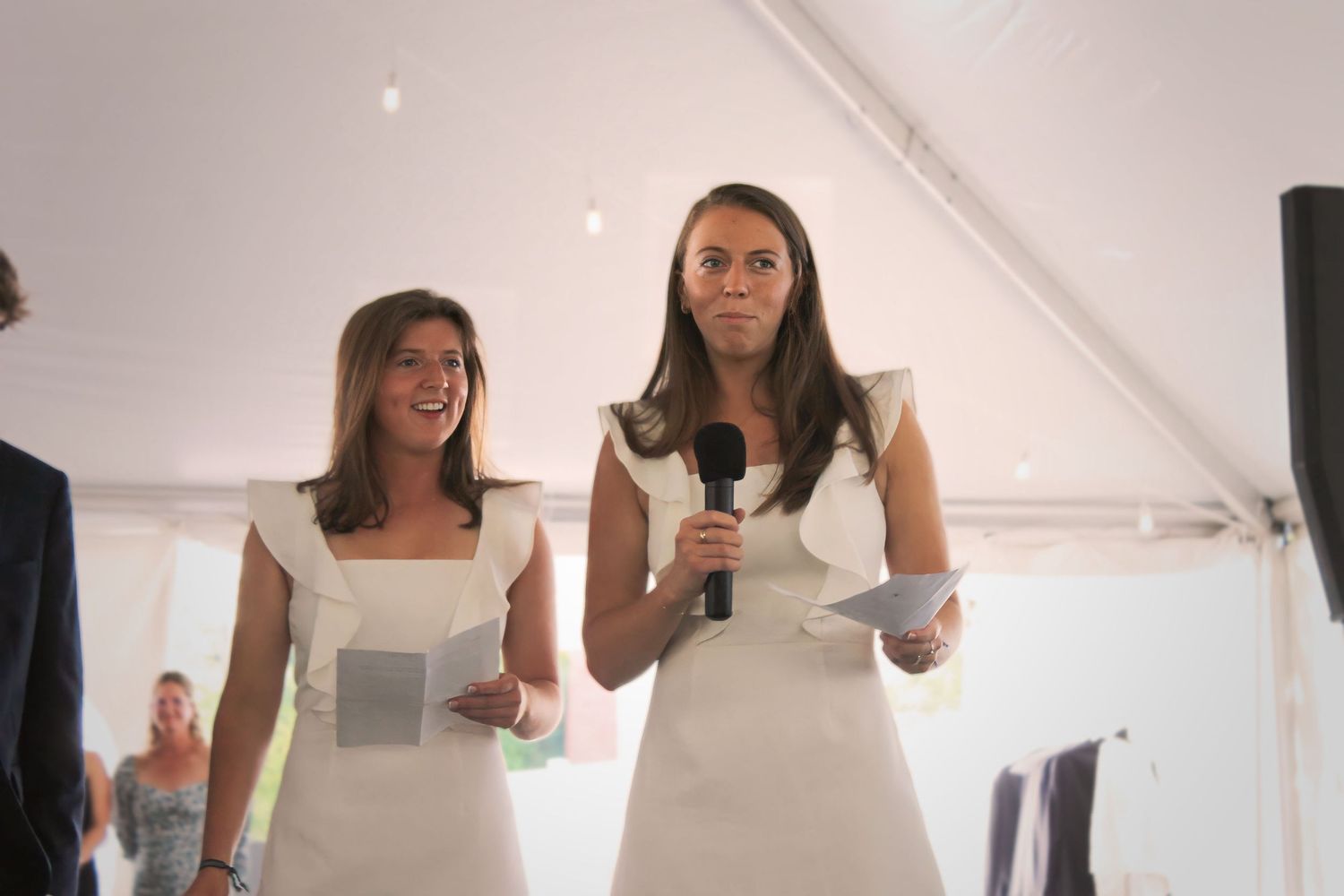 Two women in white dresses give speeches under a white tent.
