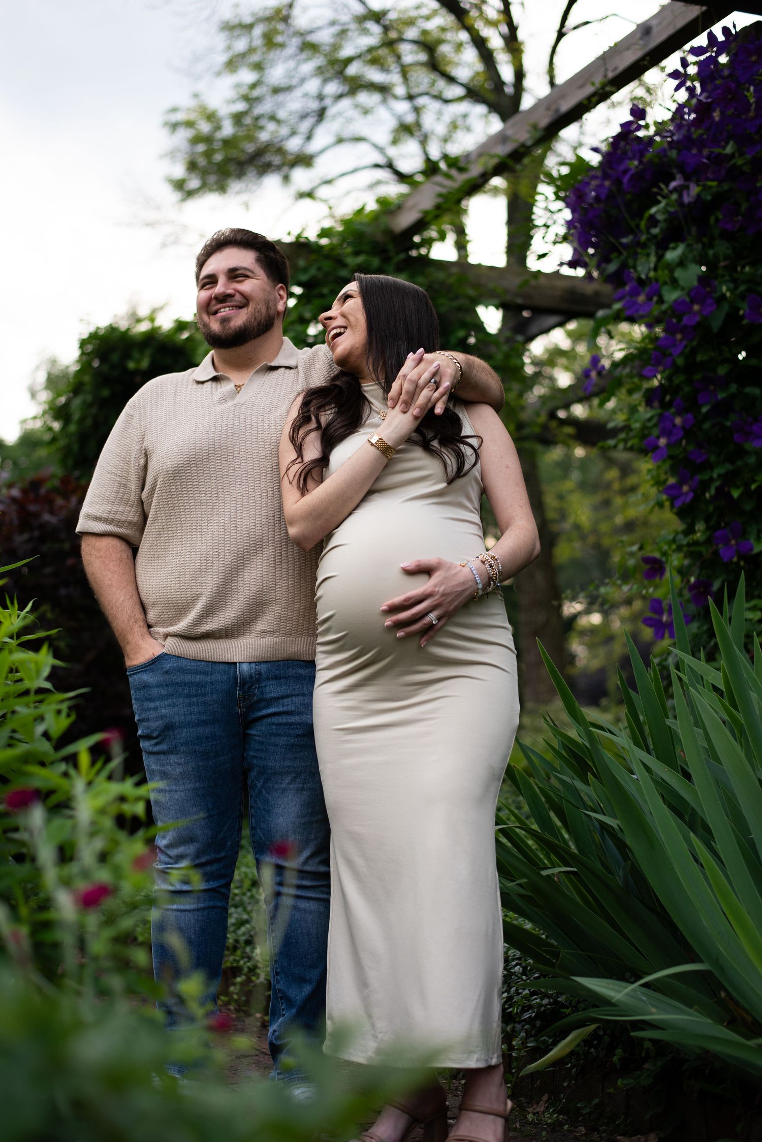Maternity photoshoot in a lush garden with purple flowers and greenery.