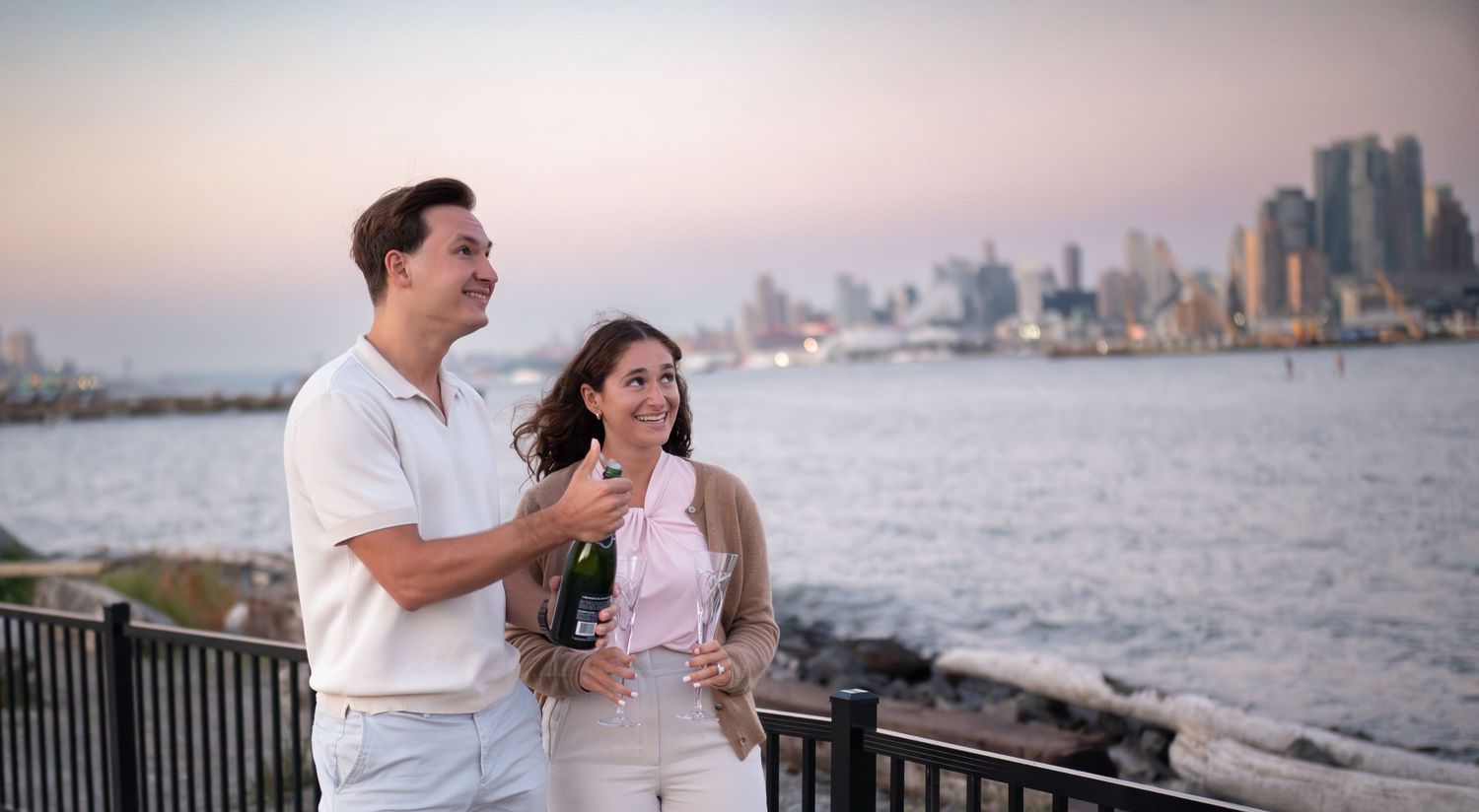 Couple shares romantic moments by the waterfront at sunset with city skyline in the background.
