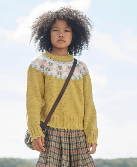 Child wearing yellow knit sweater with fair isle pattern and plaid skirt poses outdoors against cloudy sky.
