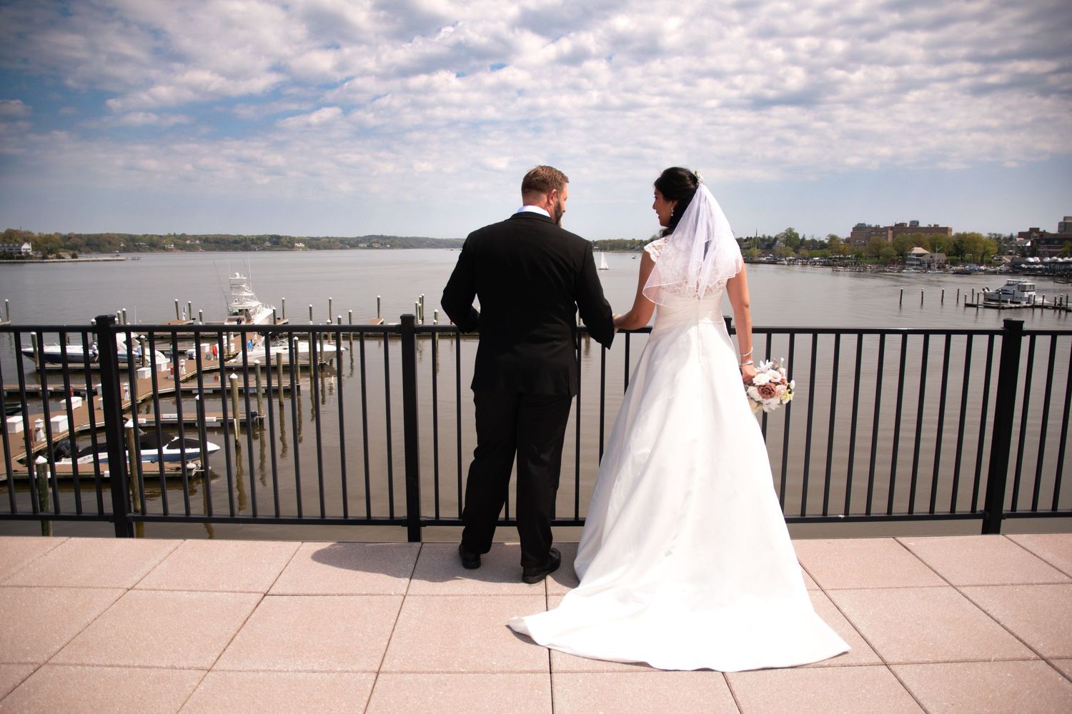 Wedding couple standing on waterfront balcony overlooking harbor view.