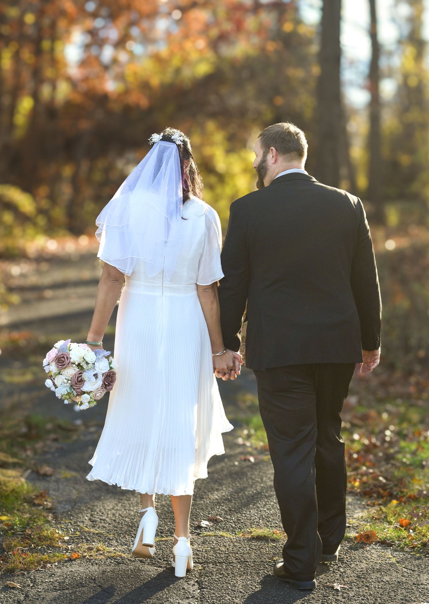Couple walks hand in hand down autumn path in formal attire.