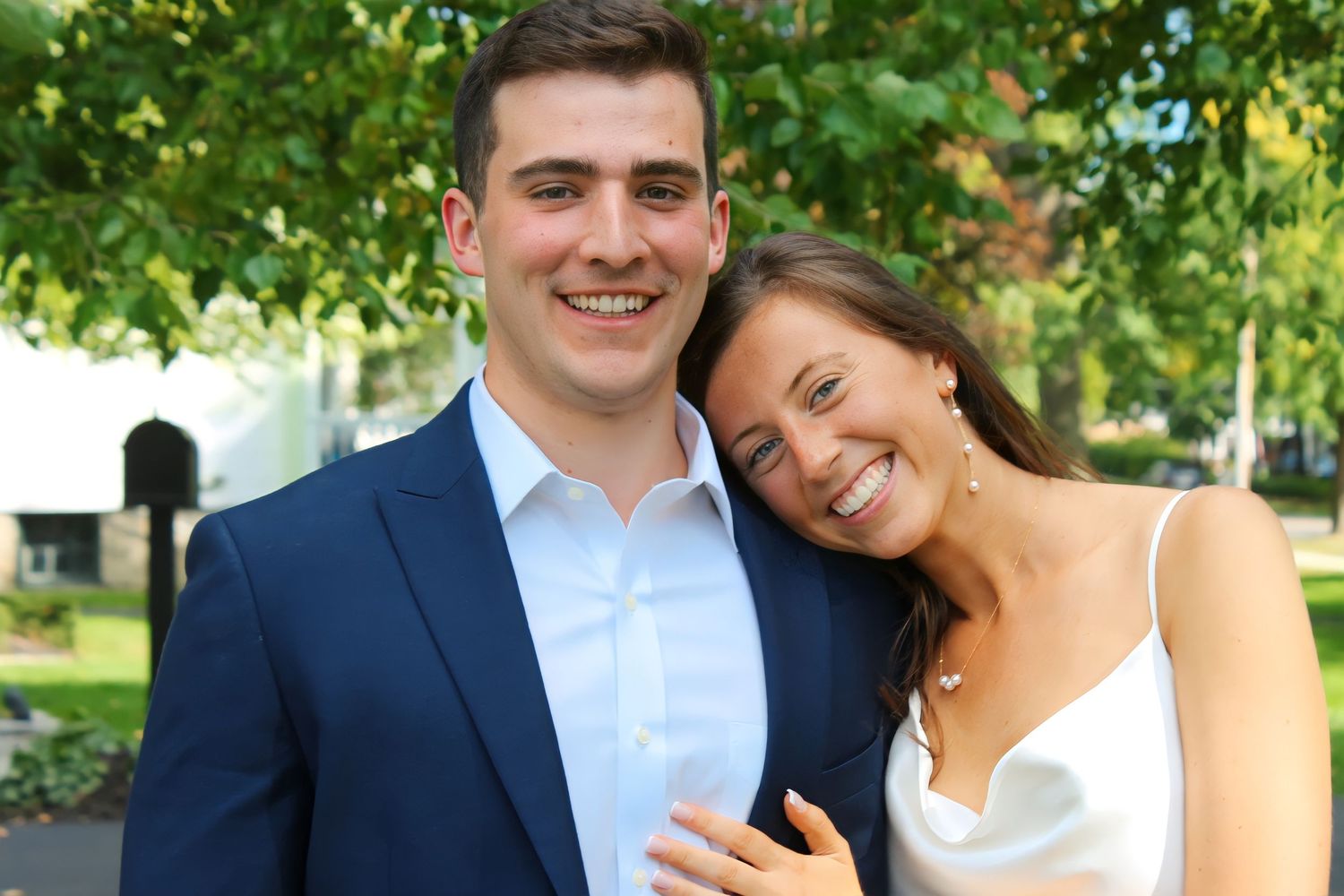 A couple poses together under green trees for engagement photos.