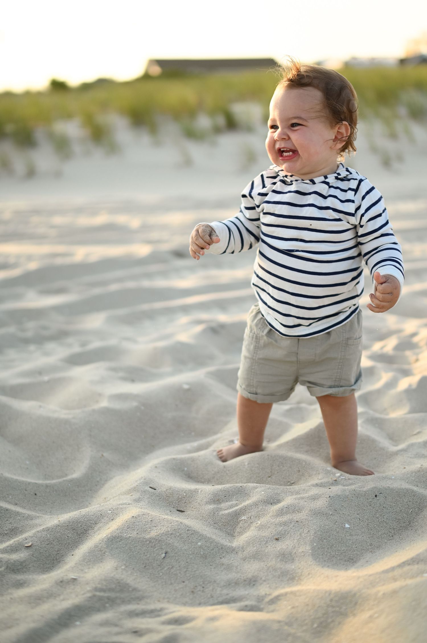 Baby in striped shirt and shorts standing on sandy beach at golden hour.