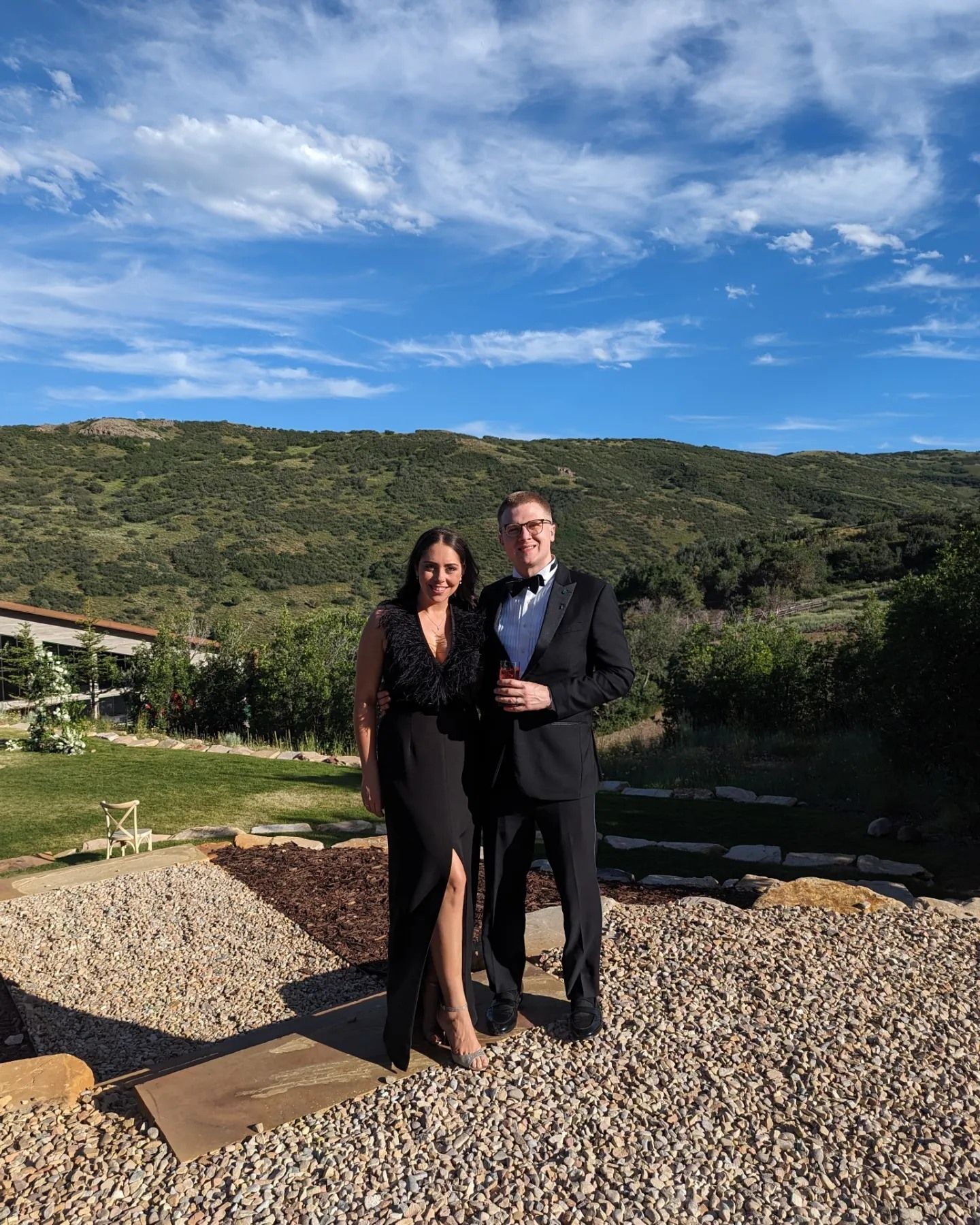 Couple in formal attire poses at sunset on a scenic overlook with mountains in the background.