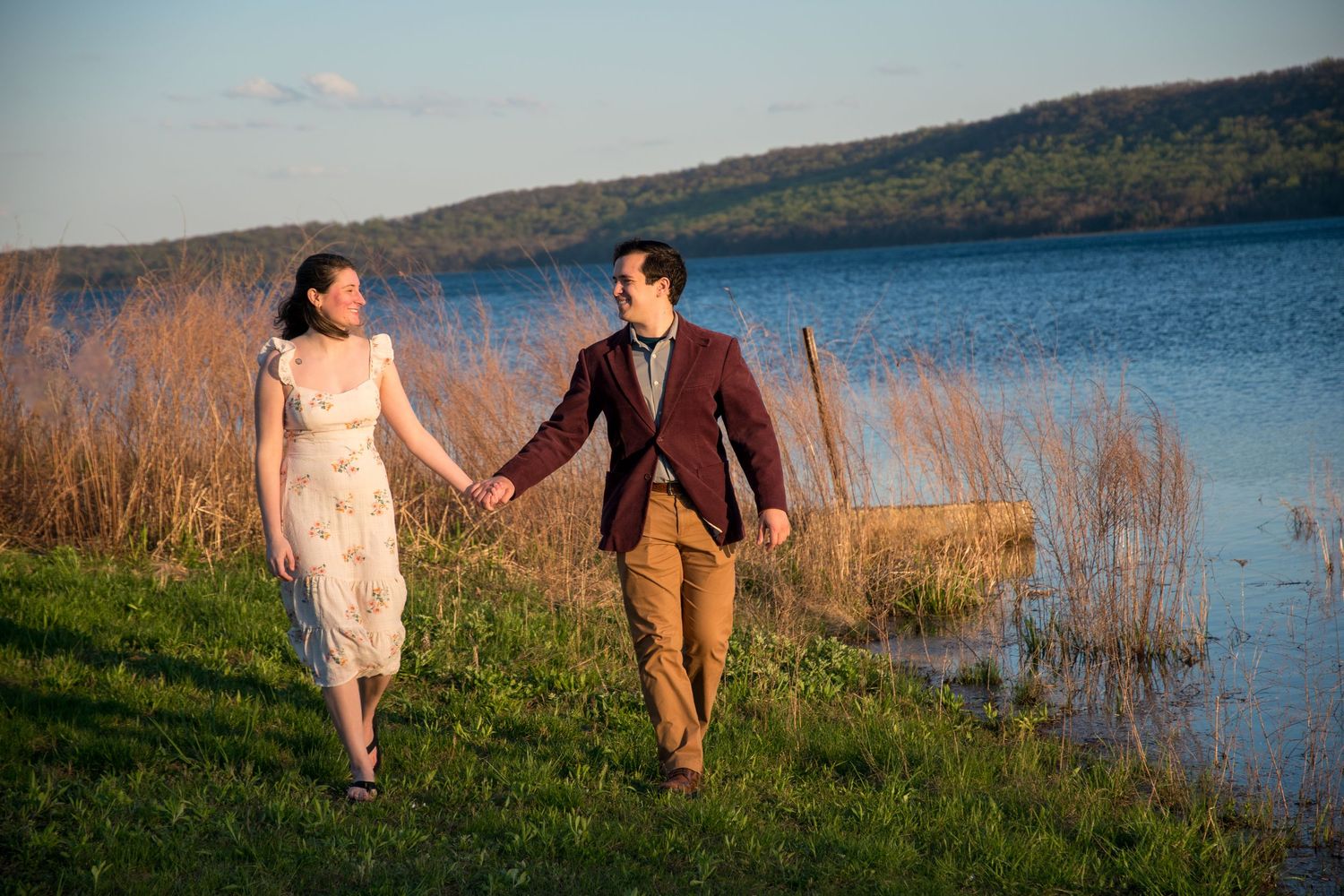 Romantic pair walks along grassy lakeside path during sunset.