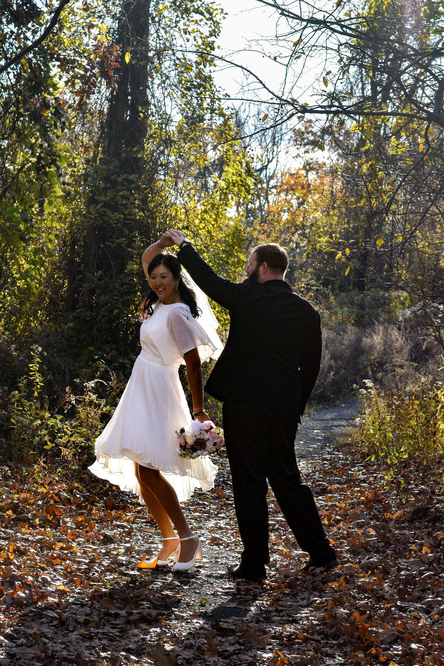 Playful couple in formal wear walks through leaf-covered forest path.