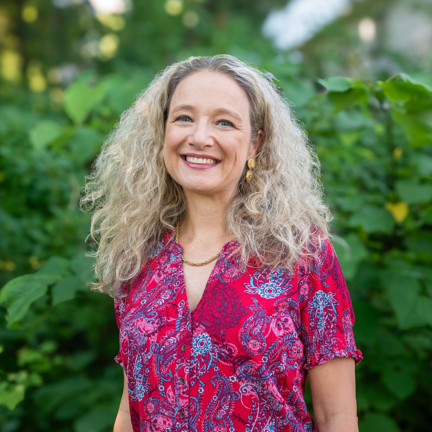 Professional outdoor portrait with red floral dress against green garden backdrop.