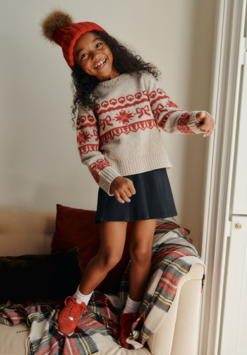 Playful child in festive red and white Fair Isle sweater and navy pants poses energetically on window ledge.