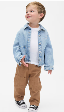 Young child wearing a light denim jacket, brown pants and black sneakers poses against white background.