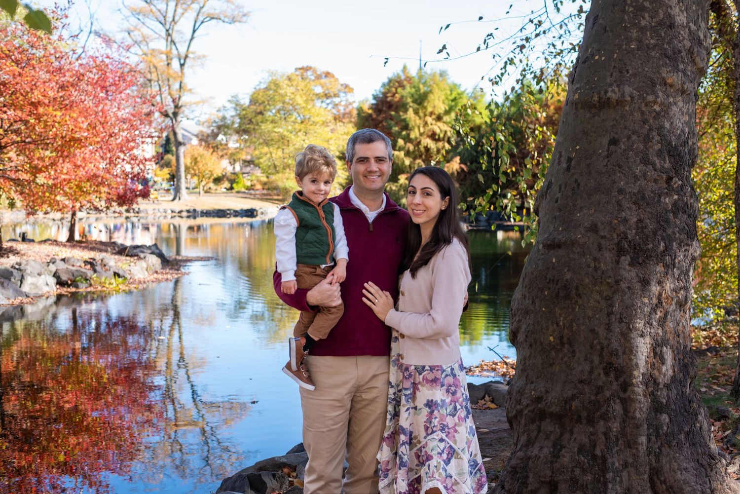 Fall family portraits near a pond with colorful autumn trees reflected in the water.
