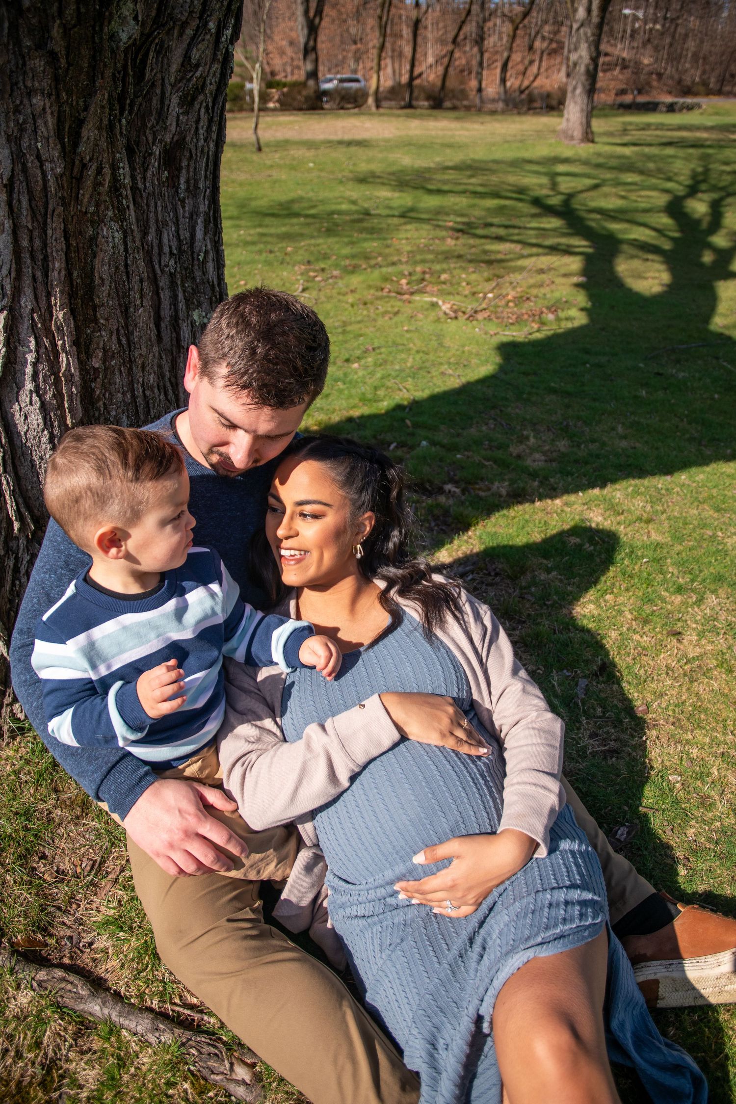 Family sitting together outdoors against large tree trunk in park setting.