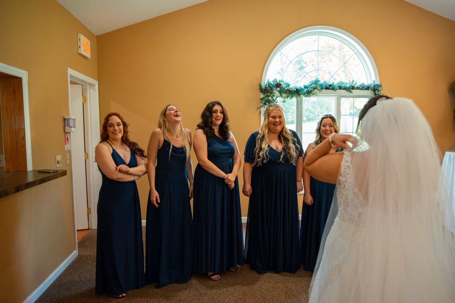 Wedding party lined up in navy blue dresses against orange wall with arched window.