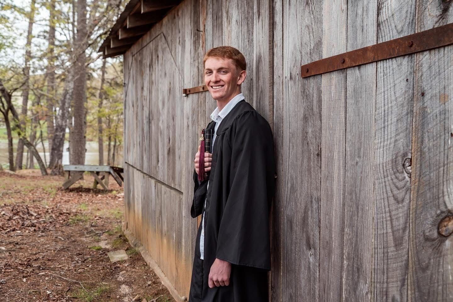 Graduate in black gown stands smiling by rustic wooden barn wall in autumn forest setting.