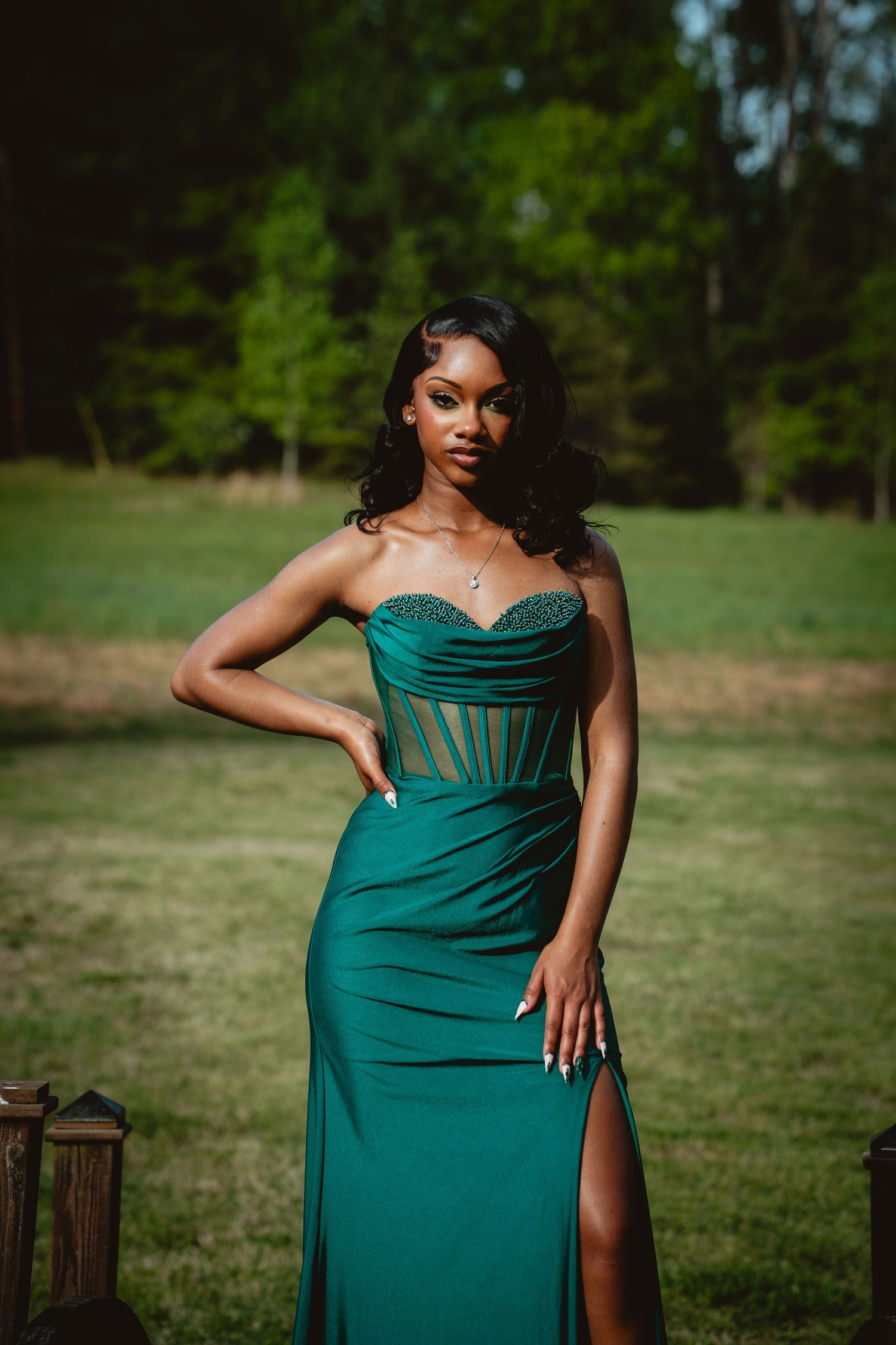 Woman in elegant emerald green evening gown with corset bodice poses outdoors in a garden setting.