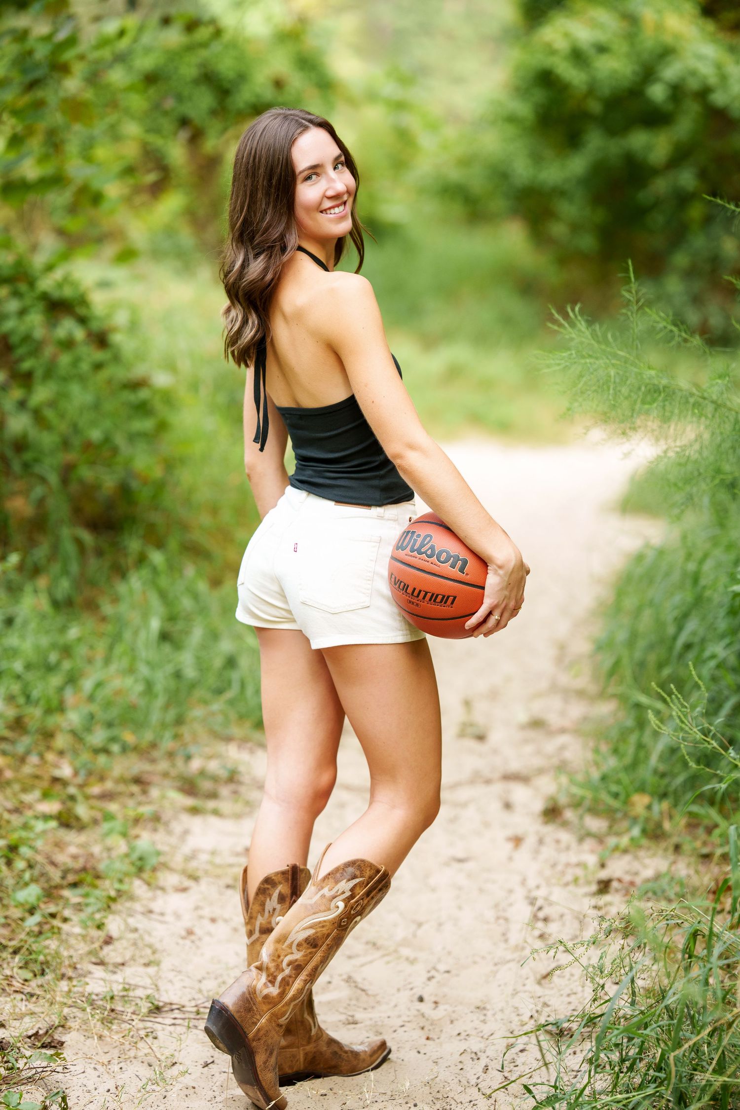 basketball senior photo in a sandy path with trees.  senior girl in boots in the woodlands with basketball.