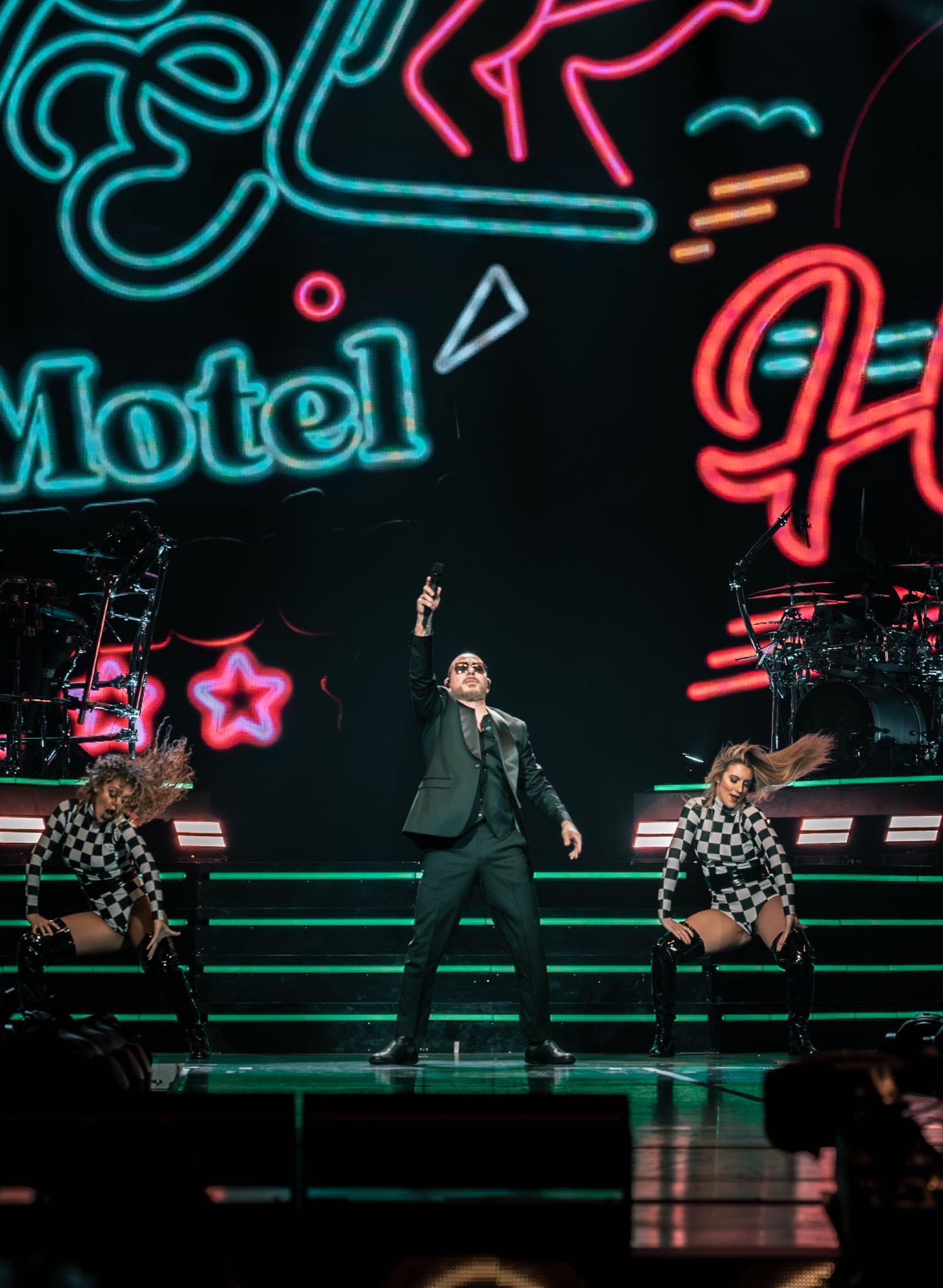 Concert stage with urban neon art backdrop and performers in coordinated black and checkered outfits.